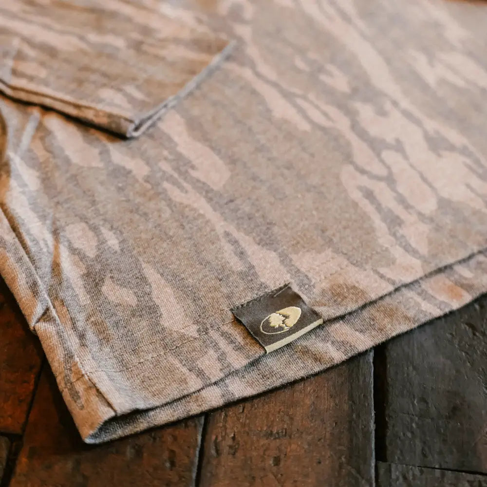 A soft, earth-toned camouflage-patterned t-shirt with a subtle texture and a small embroidered logo tag.