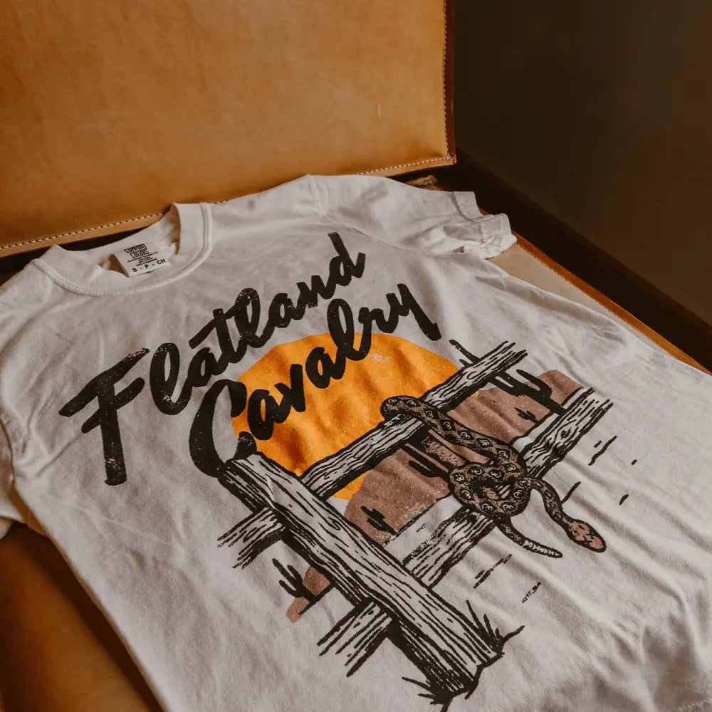White cotton t-shirt featuring a black and orange graphic of a snake coiled around a wooden fence with the text ’flatland cavalry’ in stylized lettering.