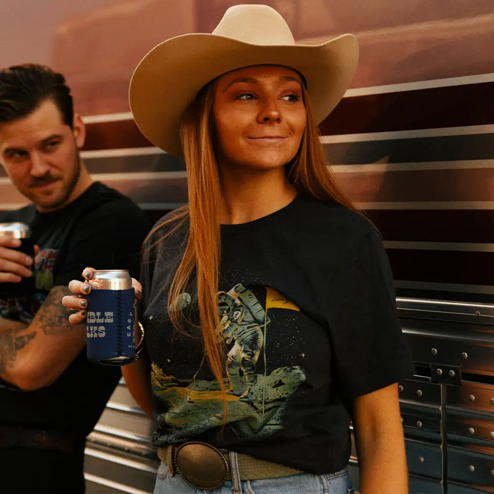 Woman wearing a cowboy hat and graphic t-shirt holding a drink can.