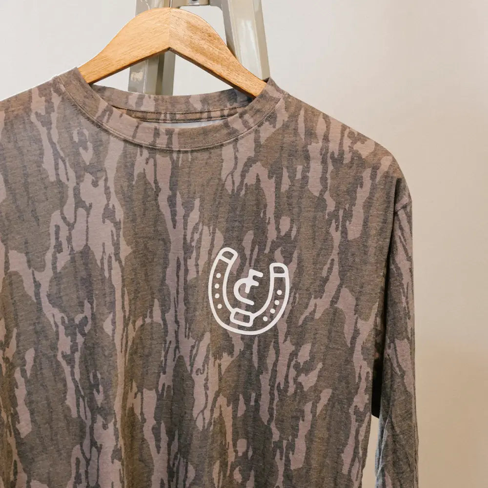 A long-sleeved shirt with a muted camouflage pattern in shades of gray and brown, featuring a white horseshoe logo on the chest.