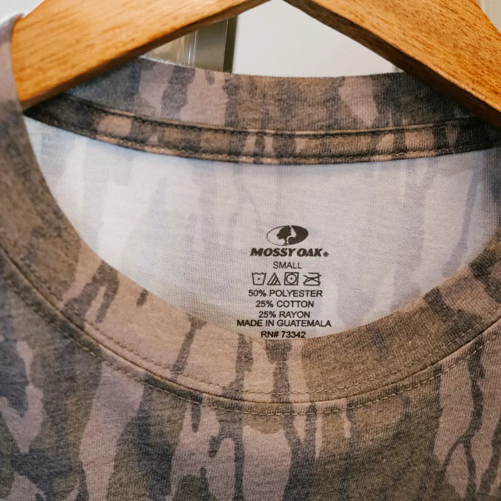 Mossy oak brand t-shirt with a camouflage pattern in shades of brown and gray, made from 50% polyester, 25% cotton, and 25% rayon.