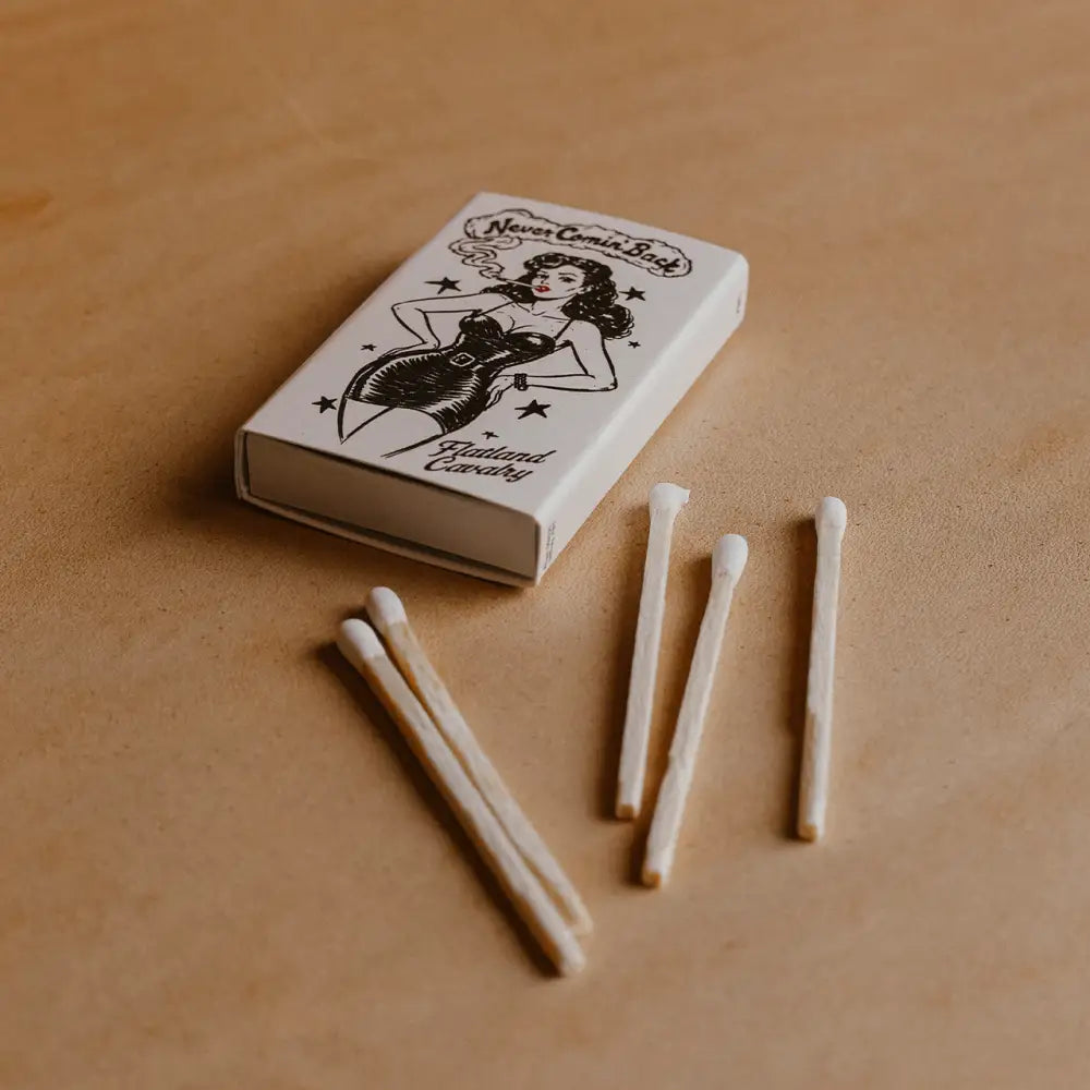 White matchbox with black ink illustration of a pin-up woman and the text ’never comin’ back’ and ’rita’s cavalry.’