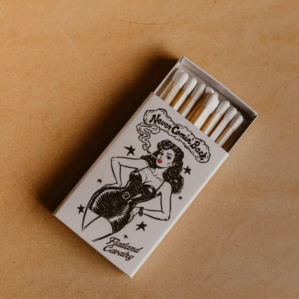 White matchbox featuring a black ink illustration of a pin-up woman smoking, with the text ’never comin’ back’ and ’thailand cavalry.’
