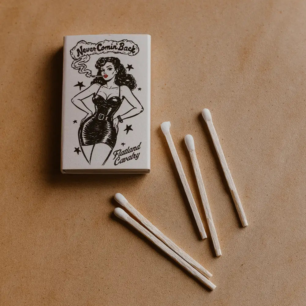 White matchbox featuring a black ink illustration of a pin-up woman smoking, labeled ’never comin’ back’ and ’flatland cavalry,’ with several wooden matches scattered nearby.