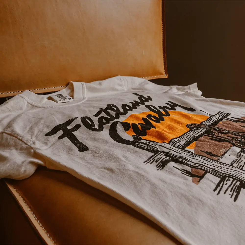 White cotton t-shirt featuring a bold black script ’flatland’ over an orange sun and rustic wooden fence graphic.