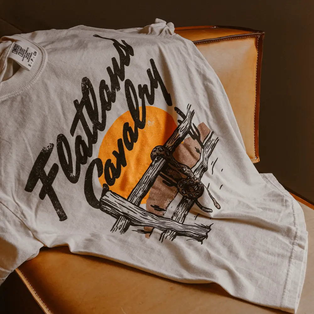 A white cotton t-shirt featuring a bold black script reading ’flatlands cavalry’ over an orange sun and rustic wooden fence graphic.
