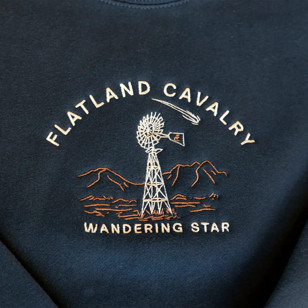Dark-colored garment with an embroidered logo featuring ’Flatland Cavalry Wandering Star’ text, a windmill, and mountain silhouettes.